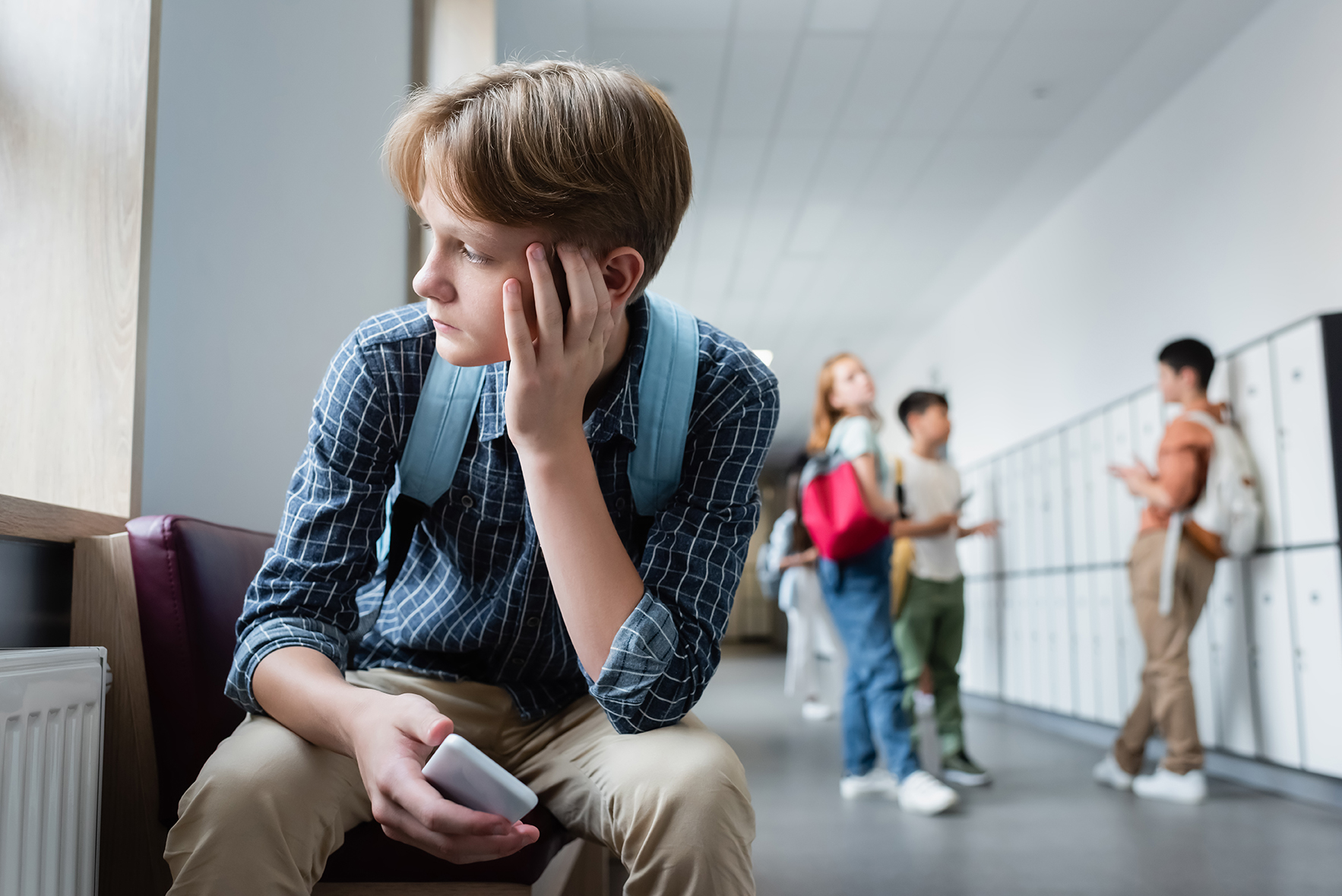 photo of boy sitting in school hallway looking glumly out window while group of schoolmates talks in the background